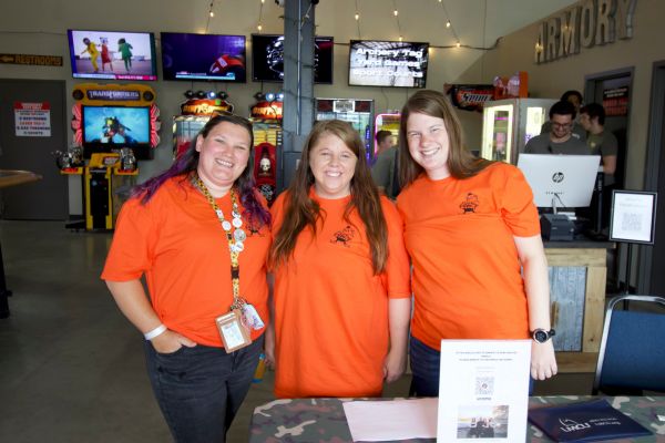 3 women in orange shirt in front of a fundraising table